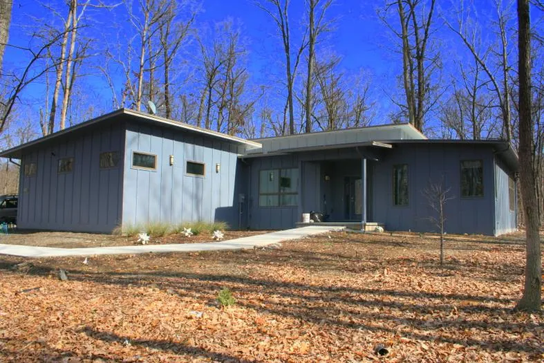 The built house: a single-story home with board-and-batten siding, shed roofs, a raised clerestory, and a covered entry, seen across a leaf-covered yard.