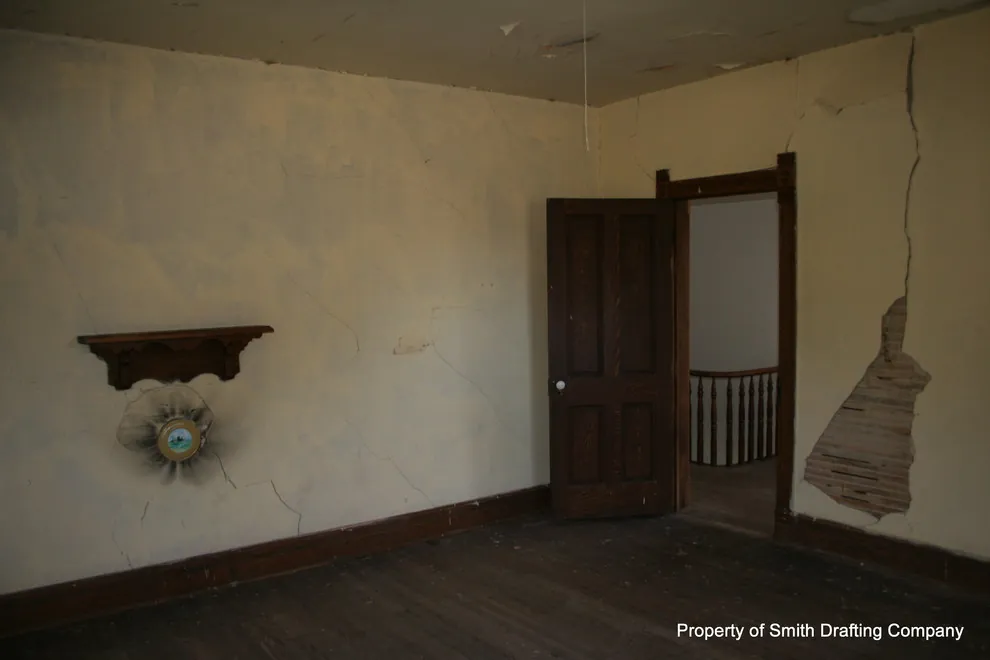 Upper room with cracked plaster, a section of missing plaster, a wall shelf, and a paneled door.