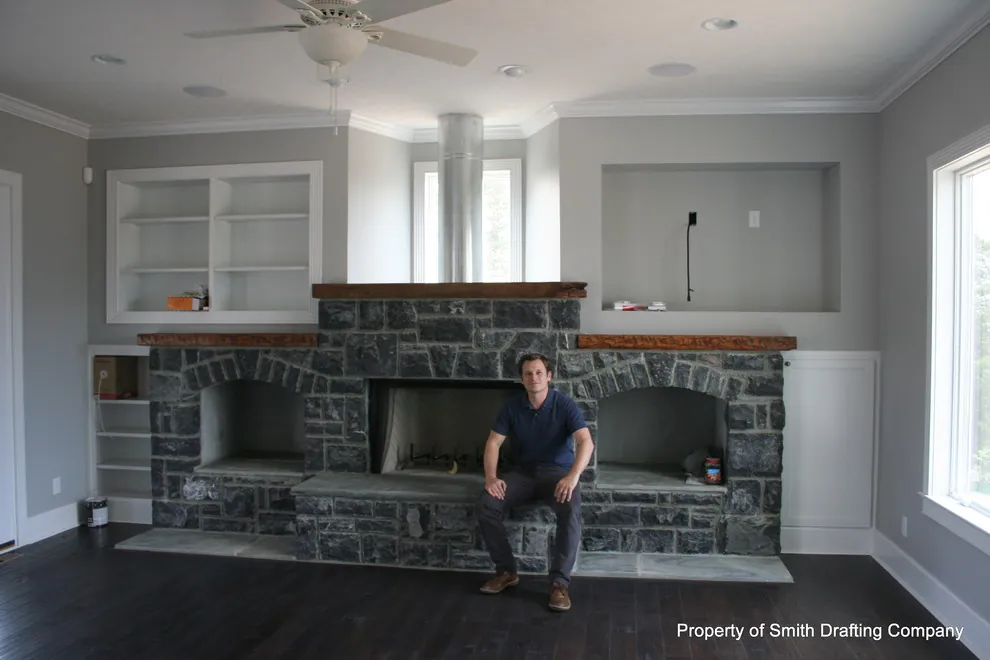 Great room with a wide stone fireplace and built-in shelves; a man sits on the hearth.