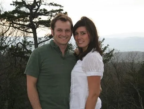 Joel and Emily Smith smiling outdoors on a ridge, with trees and hills behind them.