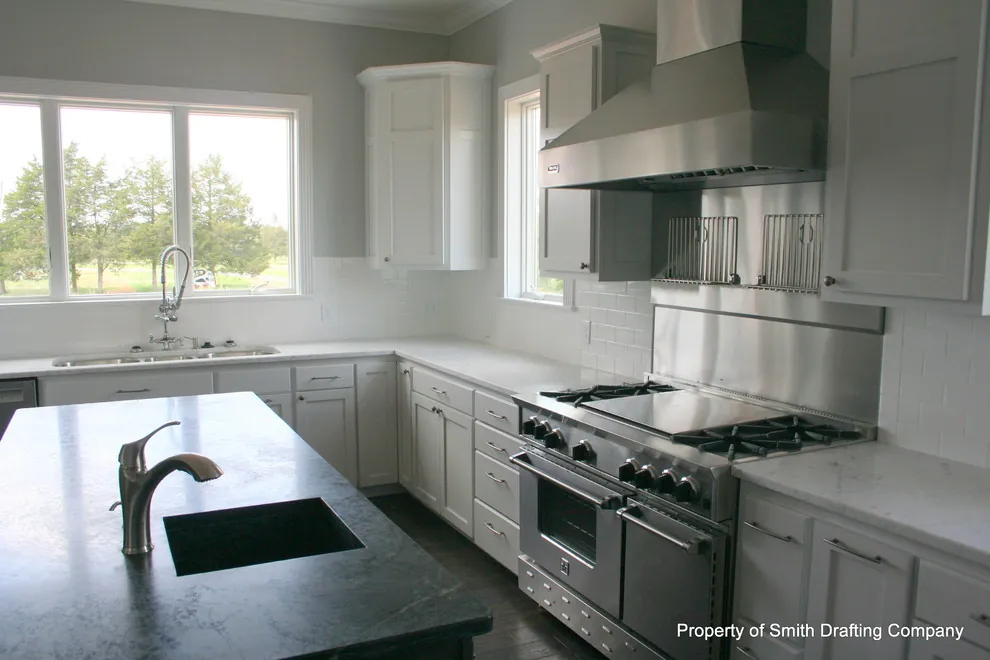White kitchen with a stainless range hood over a professional range, a dark stone island with a sink, and a wide window.